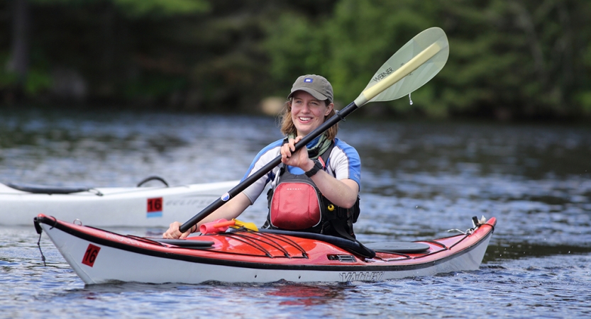 A person smiles as they paddle a kayak on calm water. 